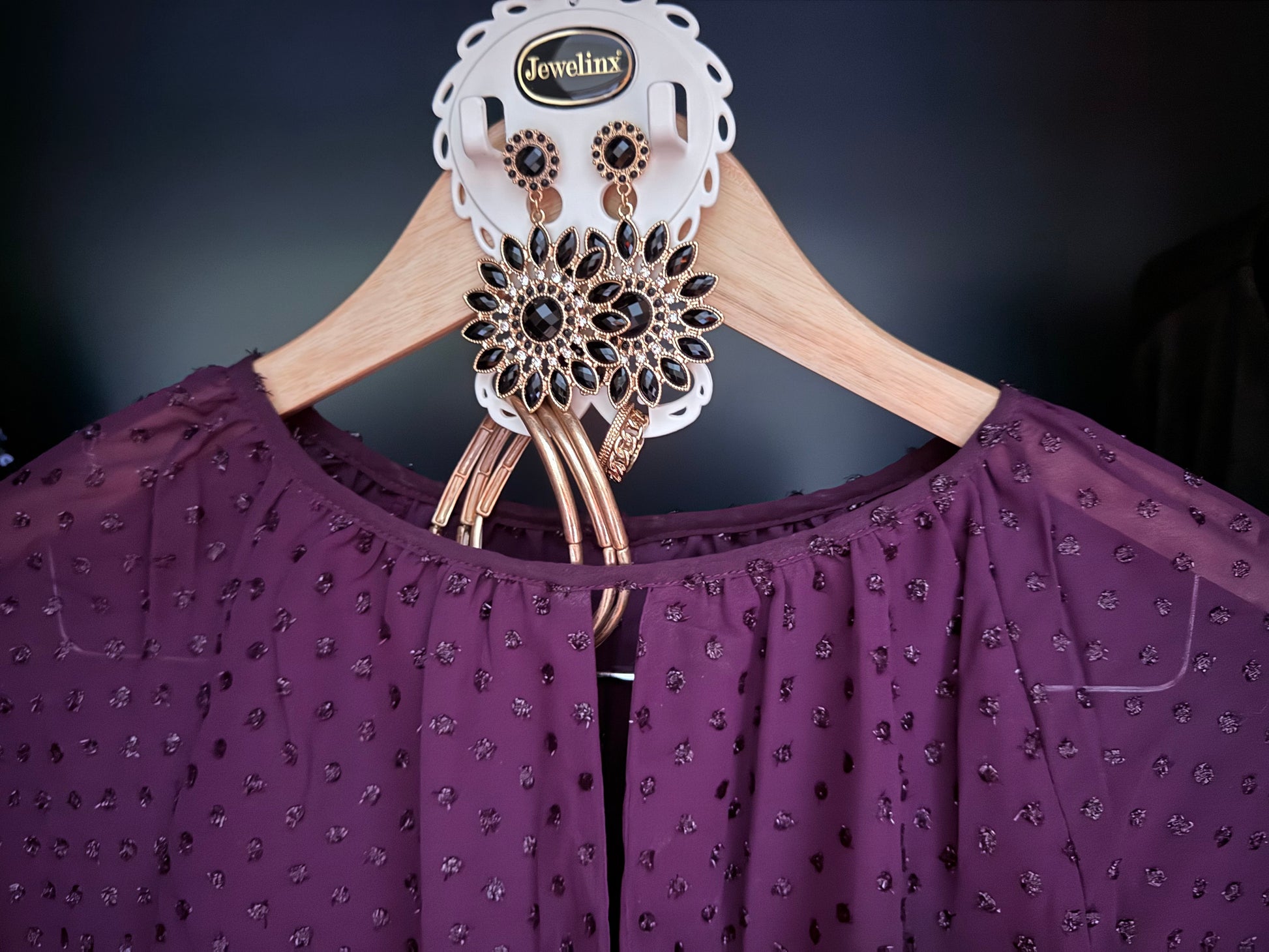 White Jewelinx hanging jewelry organizer holding black and gold statement earrings and bracelets, paired with a purple blouse